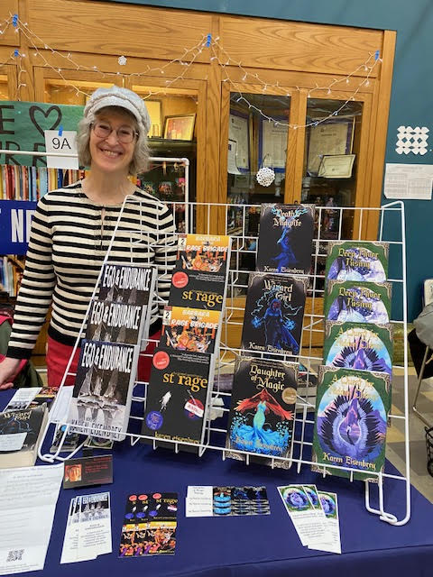 Karen smiling next to a display rack holding all of her books. She is wearing a sequined cap, striped sweater, and red pants.